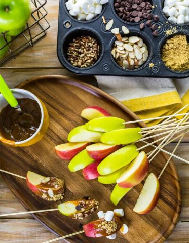 Apple slices on skewers, with a muffin tin filled with different toppins after apple is dipped in caramel. 
